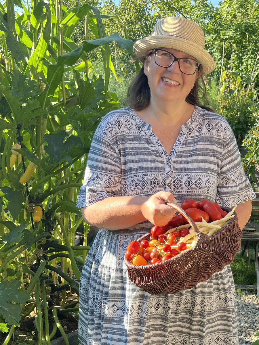 Gartentanja lacht mit Strohhut und einem Korb voller Tomaten zwischen hohen Maispflanzen in ihrem Garten am Bückeberg