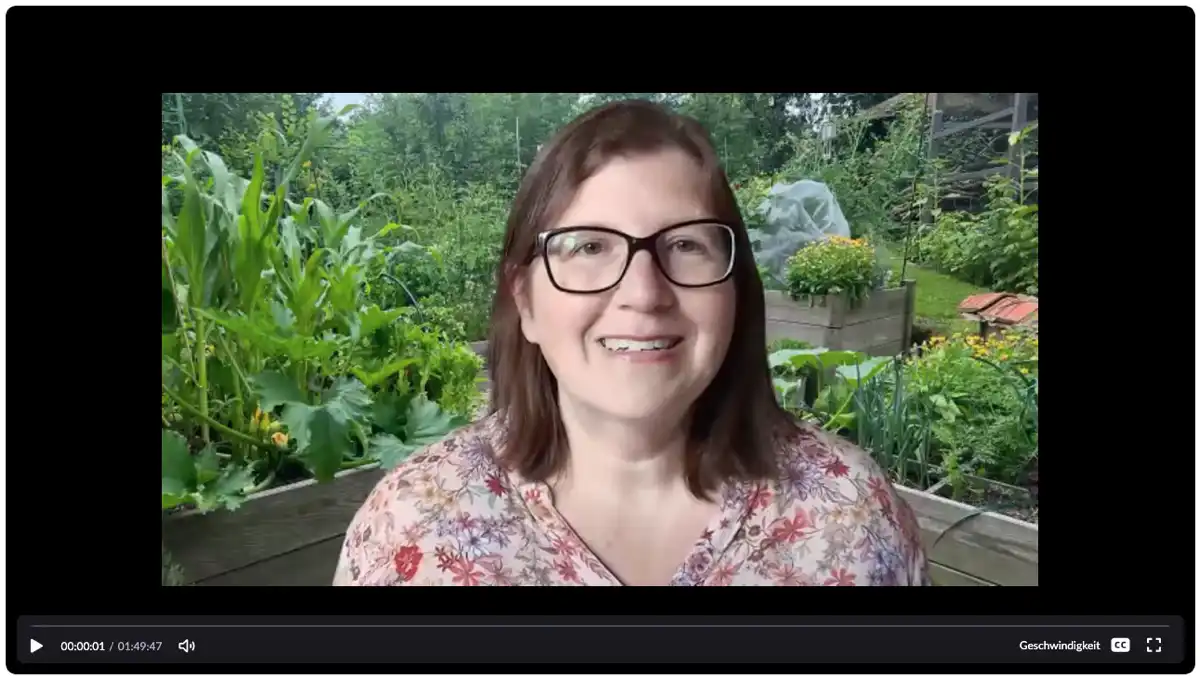 screenshot-webinar-ega-21072026 Eine Frau mit Brille und schulterlangen braunen Haaren lächelt in die Kamera. Sie trägt ein geblümtes Hemd. Im Hintergrund ist ein üppiger Garten mit grünen Pflanzen und Hochbeeten zu sehen. Am unteren Bildschirmrand sind die Videosteuerelemente zu sehen.