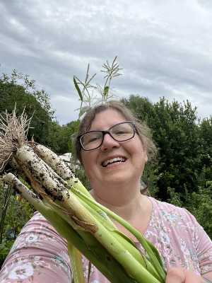 Eine lächelnde Frau mit Brille und rosa geblümtem Hemd hält frisch geerntete grüne Zwiebeln mit Wurzeln in der Hand. Sie steht im Freien in einem üppigen Garten unter einem bewölkten Himmel, mit hohen grünen Pflanzen und Bäumen im Hintergrund.